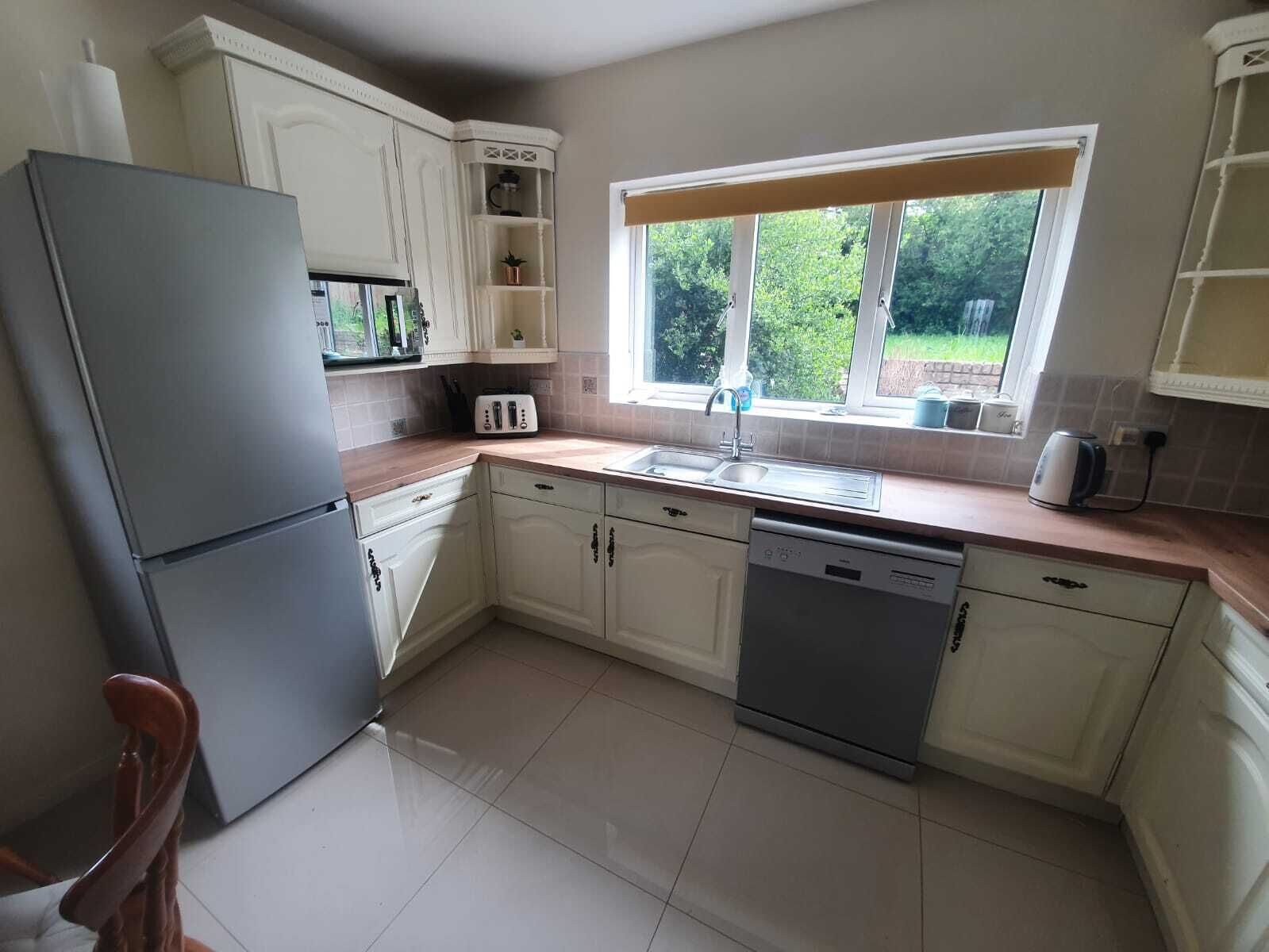 A kitchen with white cabinets, a grey refrigerator, a dishwasher, a sink, and a window with a view of greenery outside.