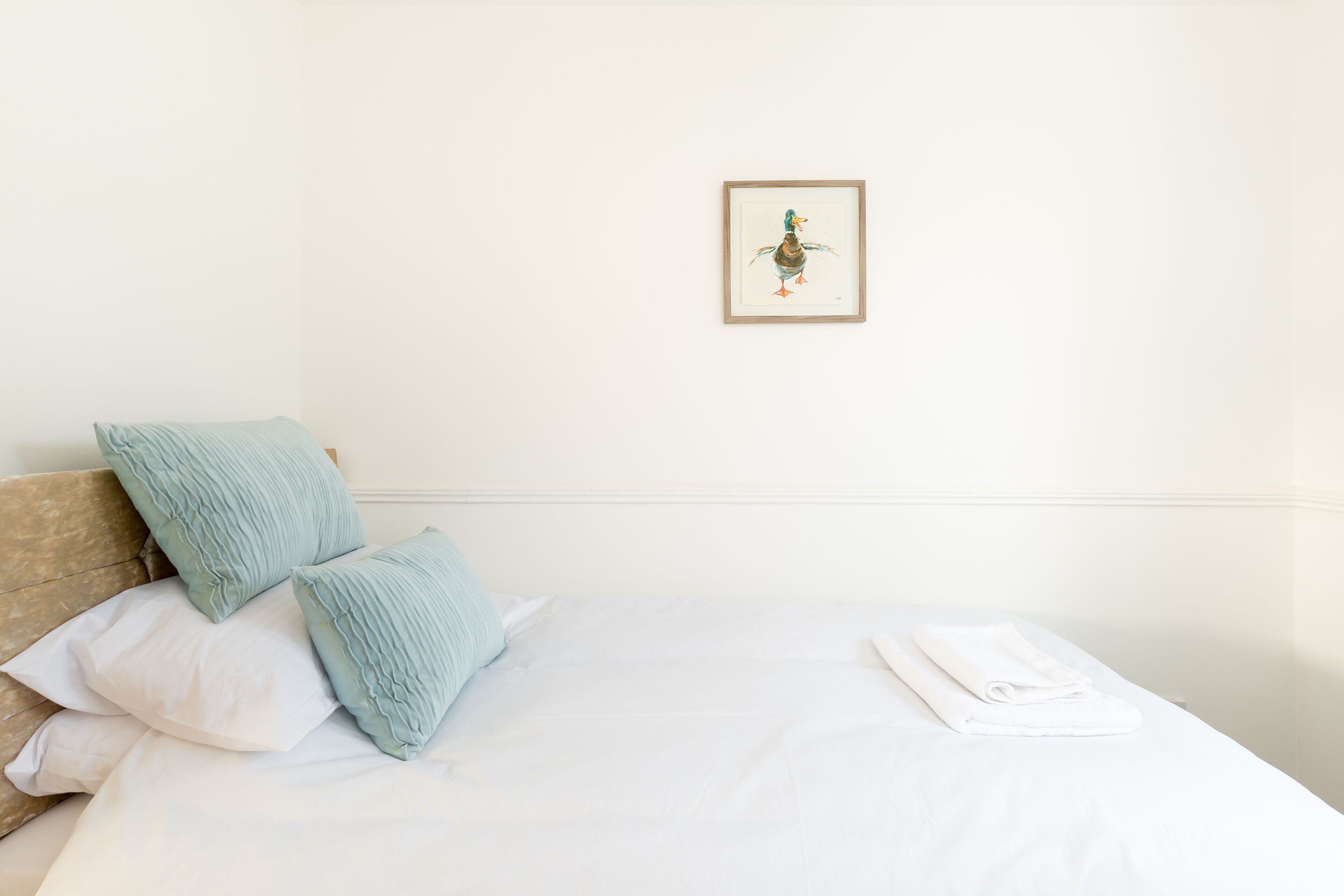 A neatly made bed with white linens and a folded towel, a pillow, and a framed picture on the wall.
