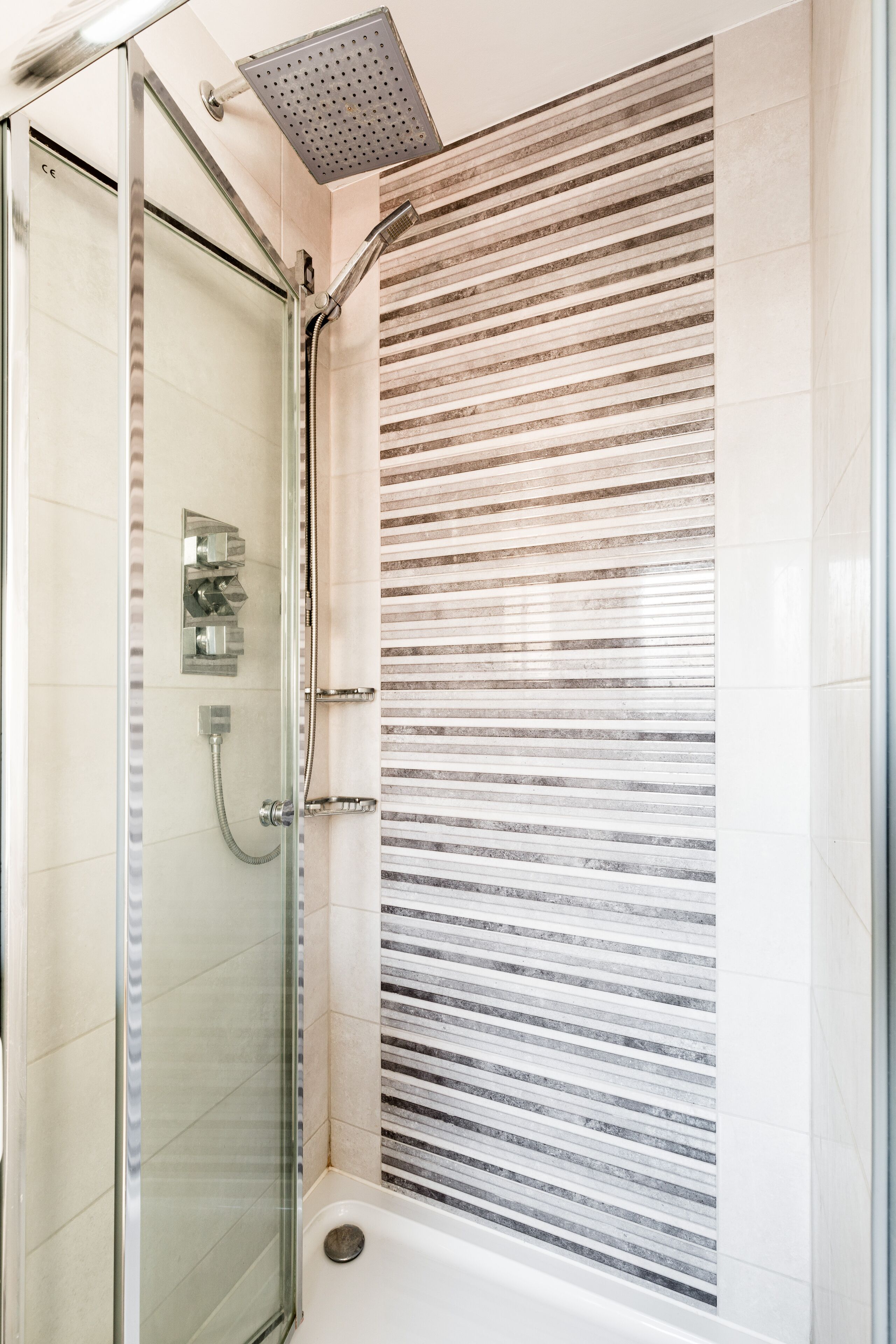 A glass-enclosed shower with a rain showerhead and a handheld showerhead, a glass door, and a tiled wall with horizontal lines.