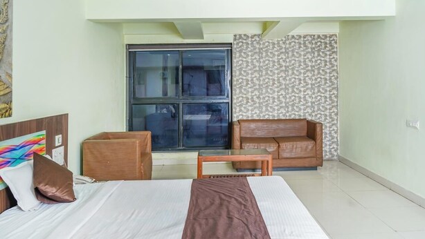 A hotel room with a bed, a wooden nightstand, a small table, a sofa, and a large window. - Lals Plaza Hotel (Bhopal)