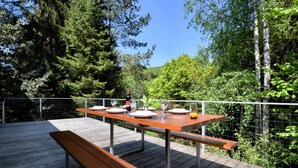 Ein Essbereich im Freien mit einer Holzterrasse, einem Metallgeländer und einem Tisch für zwei Personen, gedeckt mit Weingläsern und einer Flasche Wein.