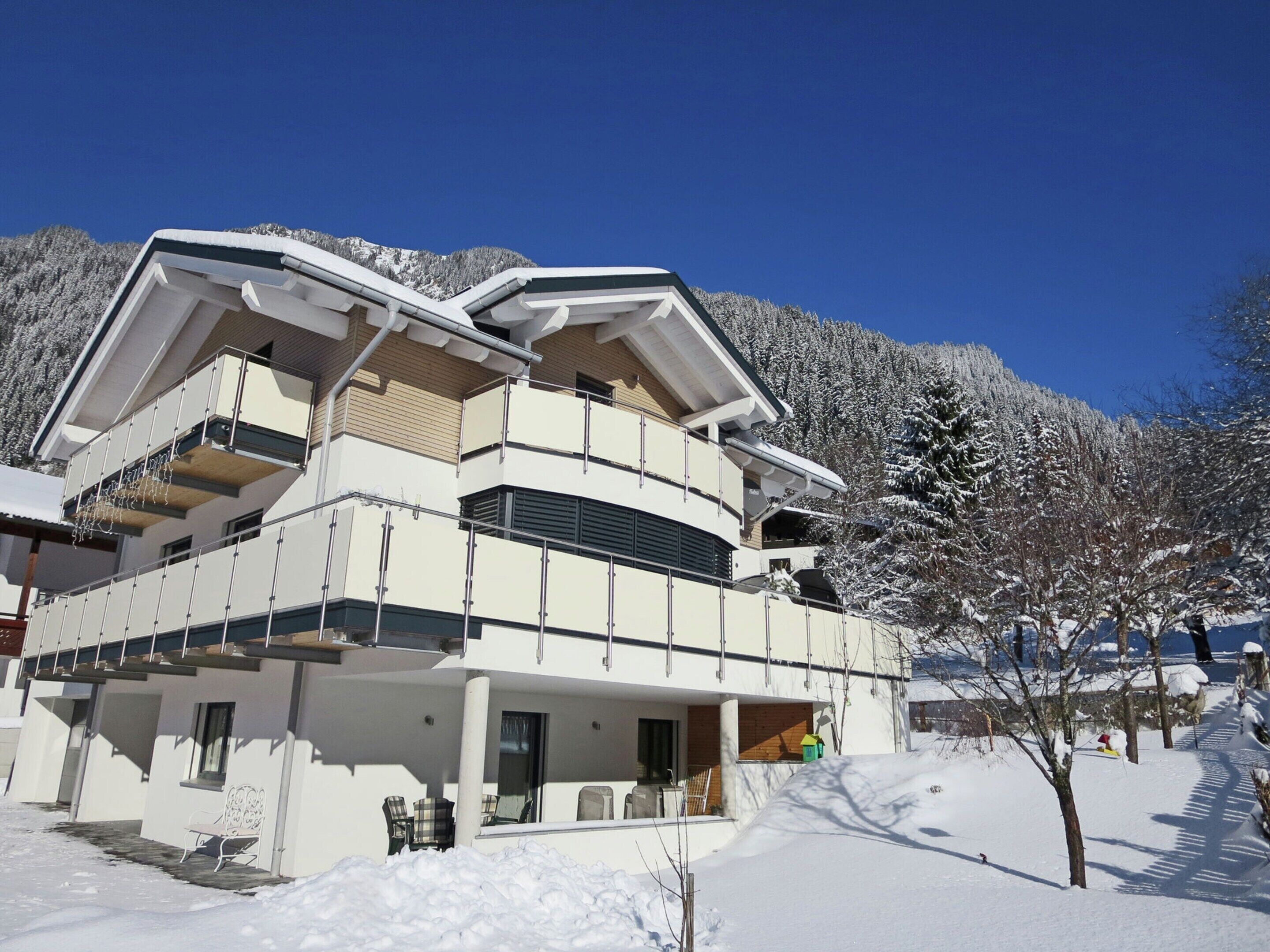 A modern multi-story building with balconies, a covered outdoor seating area, and a snowy mountainous backdrop.