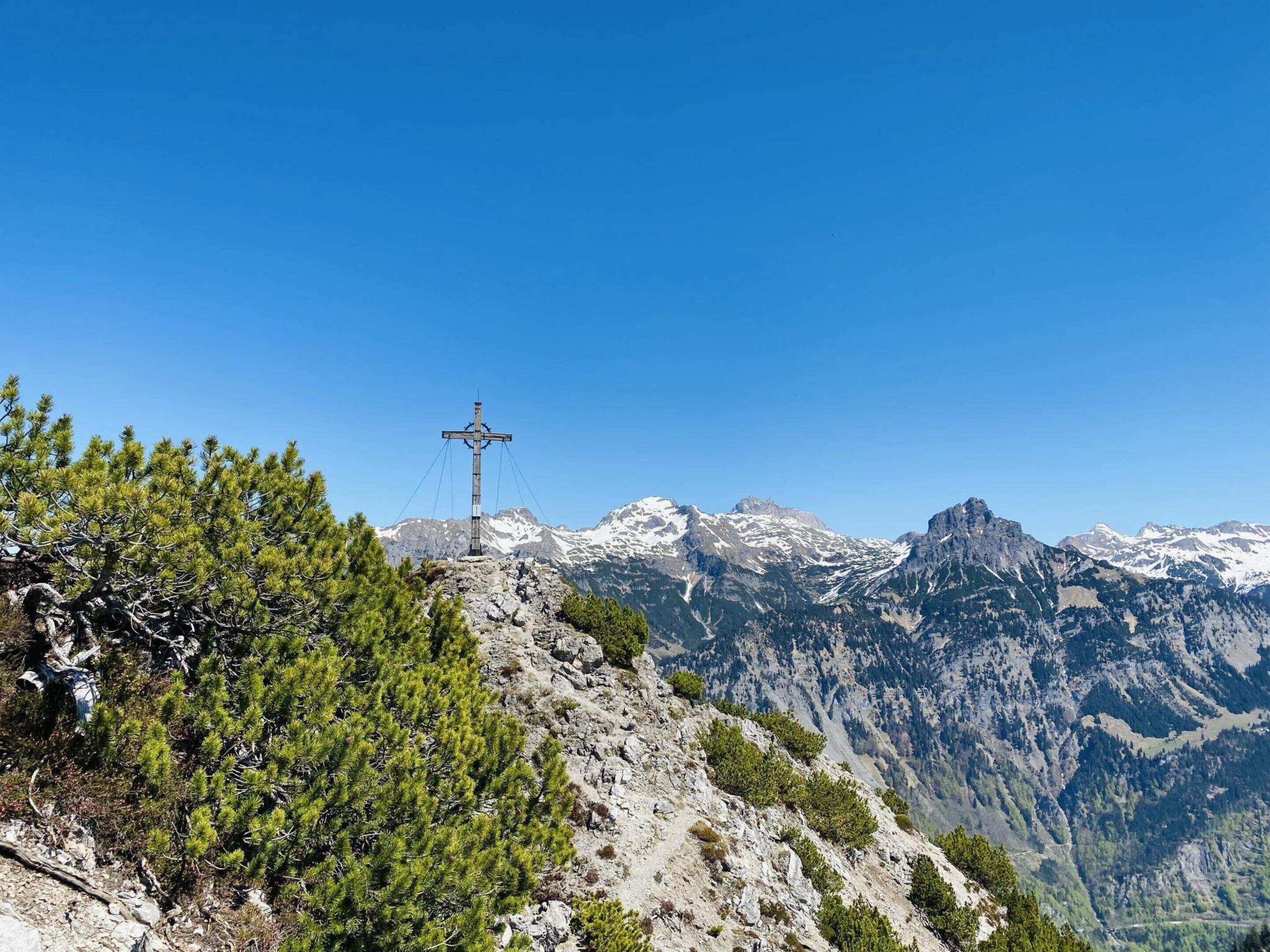 A cross on a mountain peak with snow-capped mountains in the background.