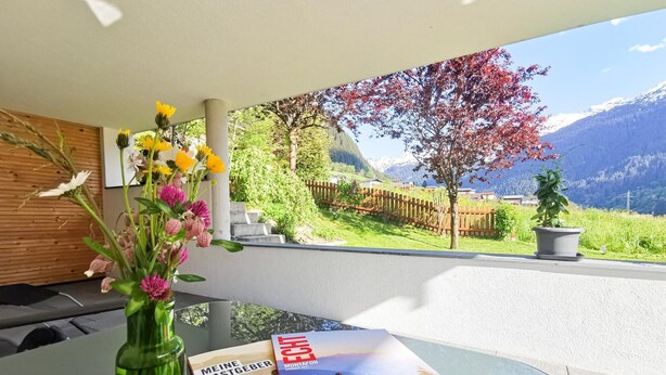A table with a vase of flowers and a magazine on a balcony overlooking a mountainous landscape.