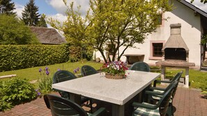 A patio with a table and chairs, a garden with flowers, and a building with a chimney. - Holiday Home in Butgenbach Near the Lake (Buetgenbach)