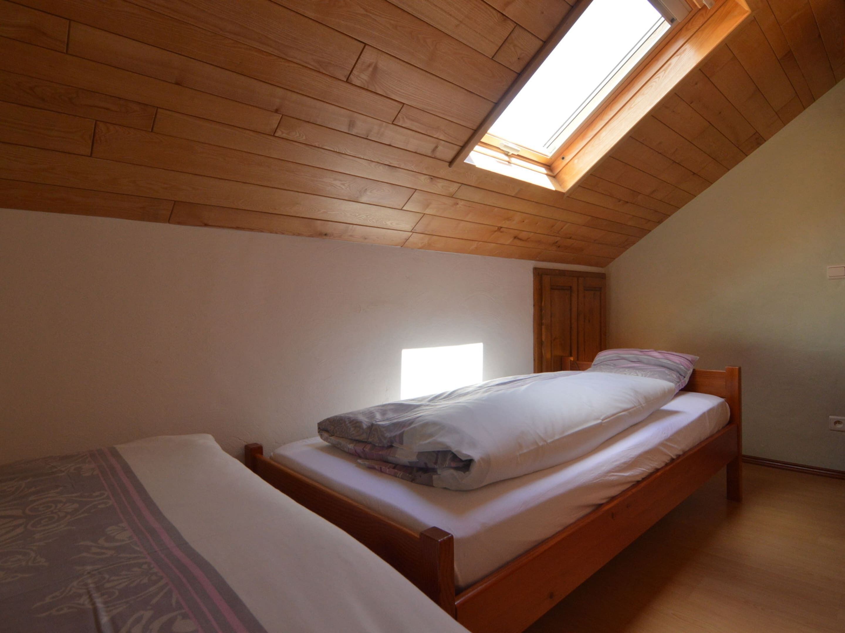 Un alojamiento pequeño y de una sola habitación con techo inclinado y tragaluz. Hay una cama con estructura de madera y ropa de cama blanca, y otra cama más pequeña con manta estampada.