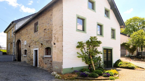 A two-story building with a stone facade and a white upper section. There are multiple windows with green frames, and a small garden with various plants and flowers. - Holiday Home in Butgenbach Near the Lake (Buetgenbach)