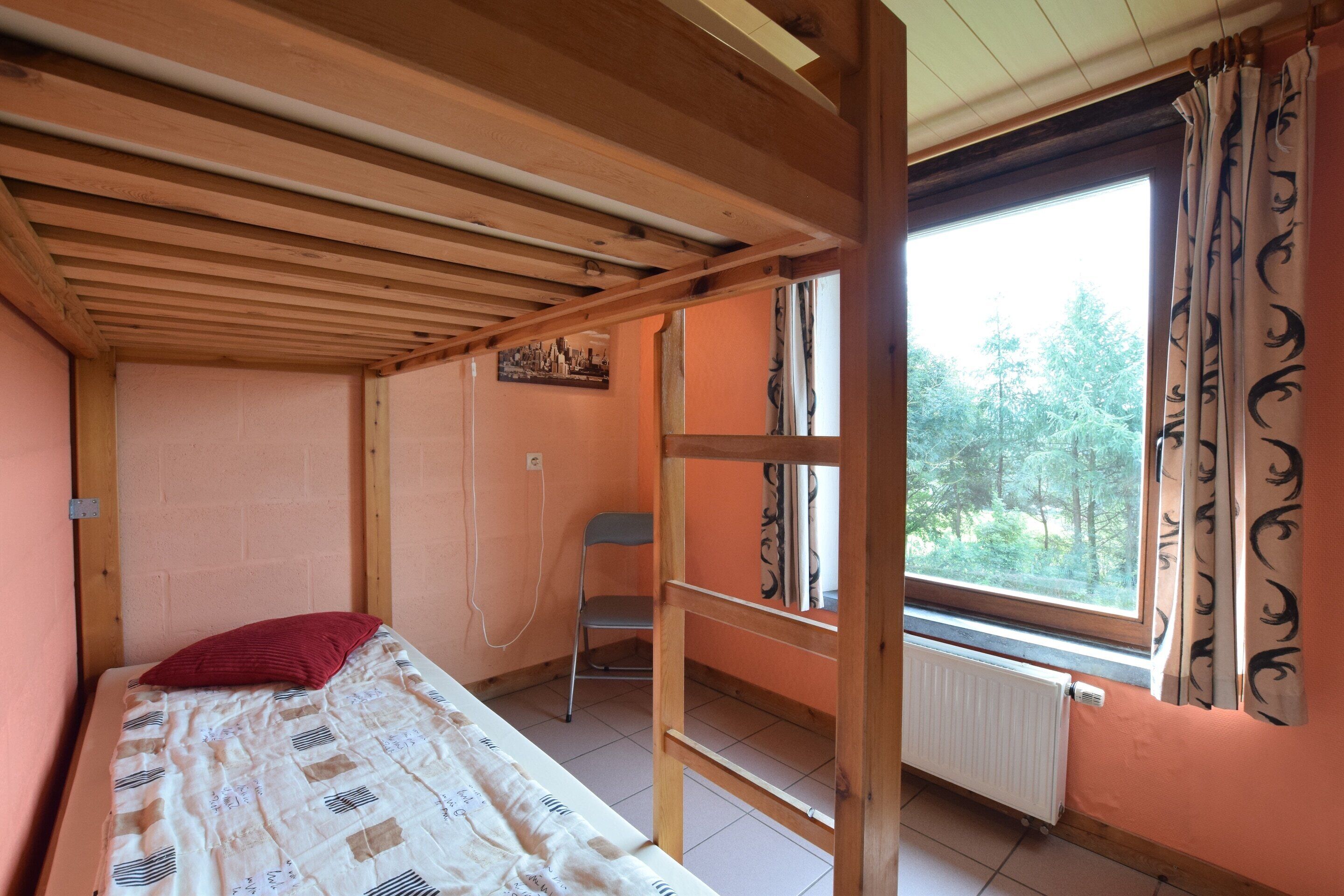 Ein Einzelbett mit rotem Kopfkissen, ein Sessel, ein Fenster mit Vorhängen und eine Heizung.