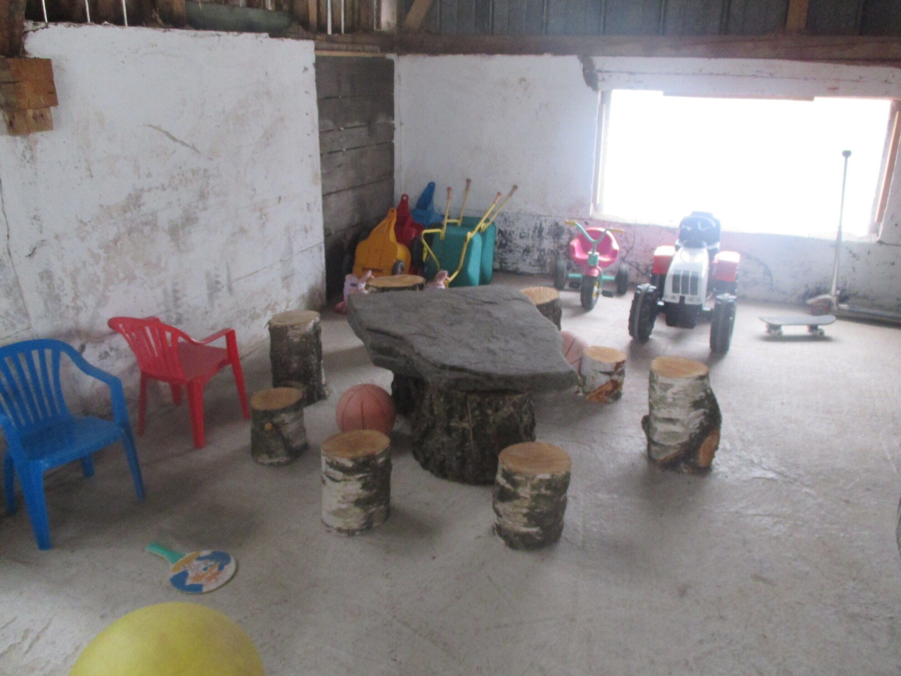 A rustic room with a stone table surrounded by wooden stools, plastic chairs, and children's toys.