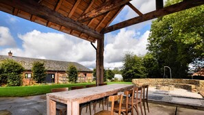 An outdoor wooden table and chairs under a covered area with a stone wall and a stone building in the background. - Pleasant Holiday Home in Erezée Ardennes with Hot Tub, Sauna (Erezee)