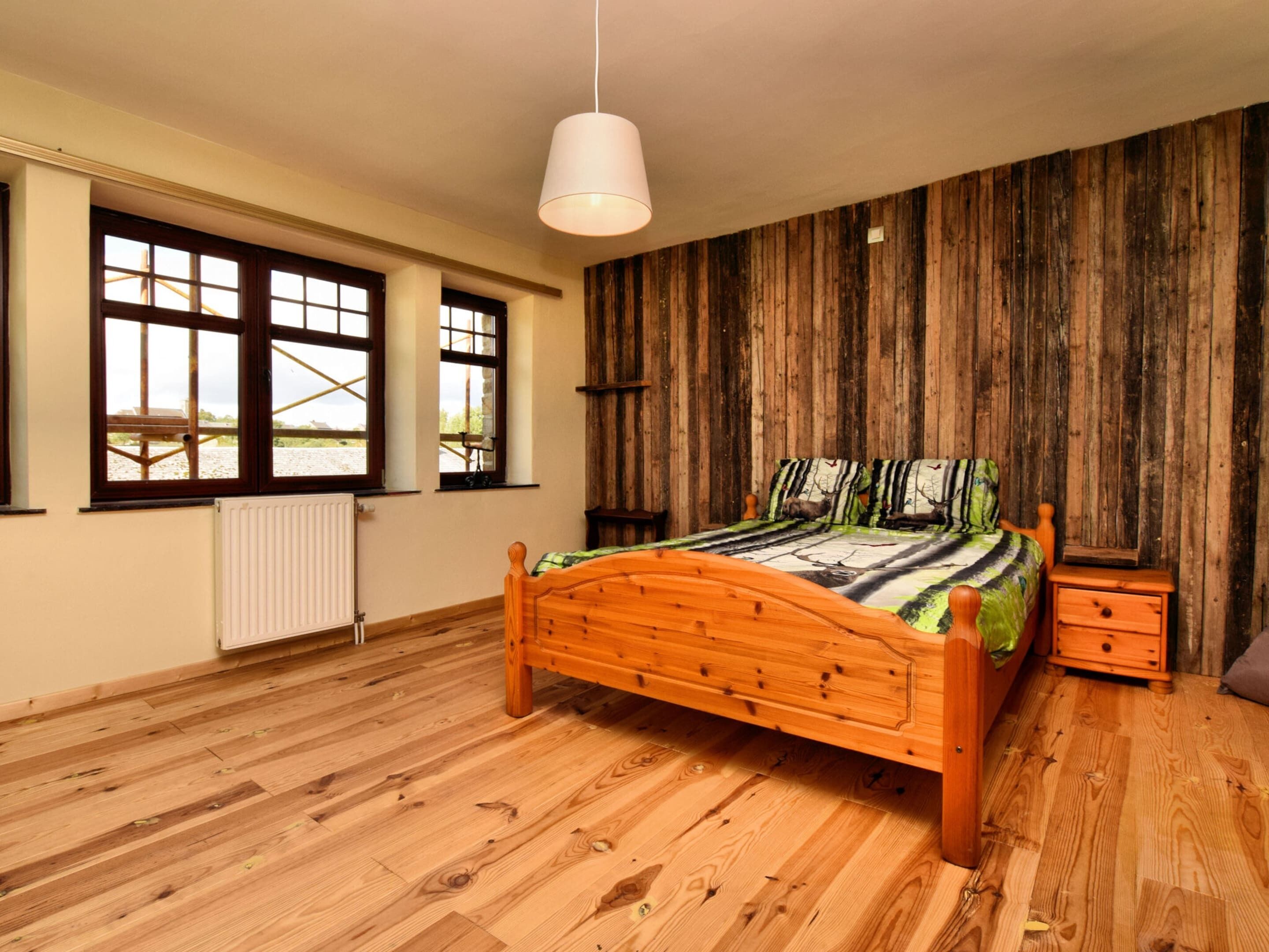 A bedroom with a wooden bed, a nightstand, a radiator, and large windows with a view.