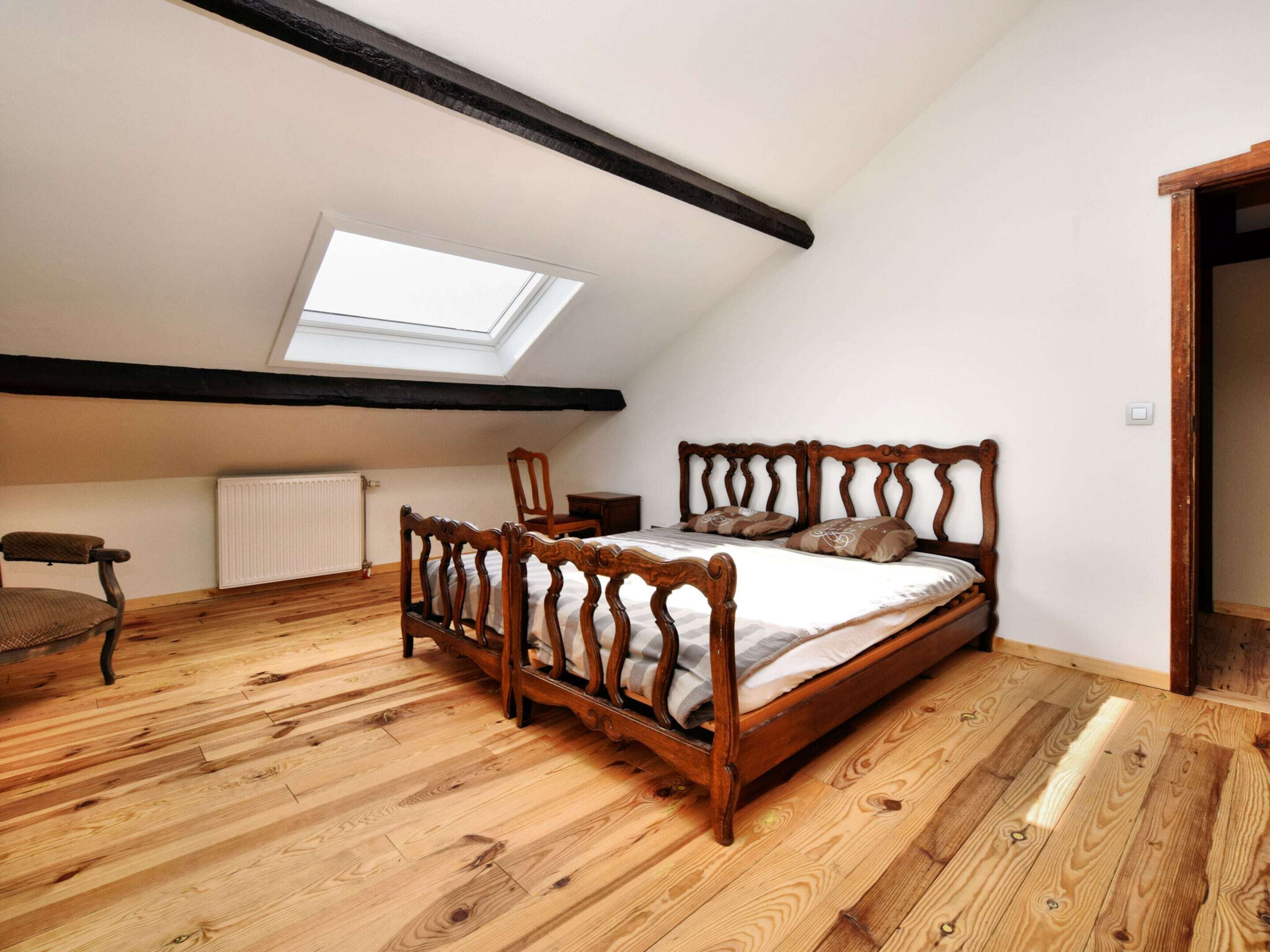 A bedroom with a sloped ceiling, wooden flooring, a bed with a wooden headboard, a chair, and a skylight.