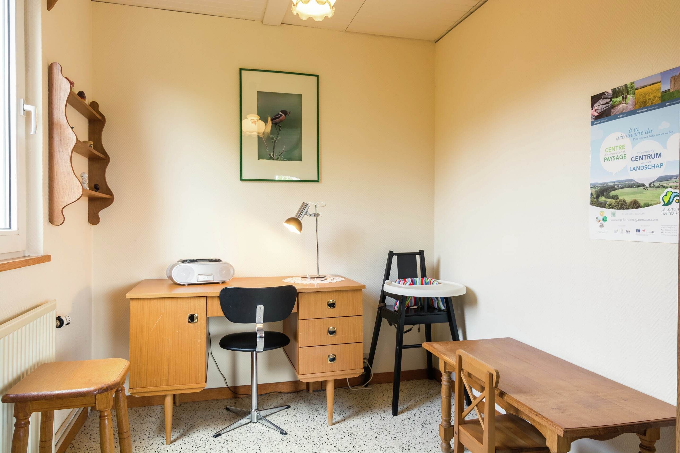 A room with a wooden desk, a chair, a small table, a radiator, a framed picture on the wall, and a poster.