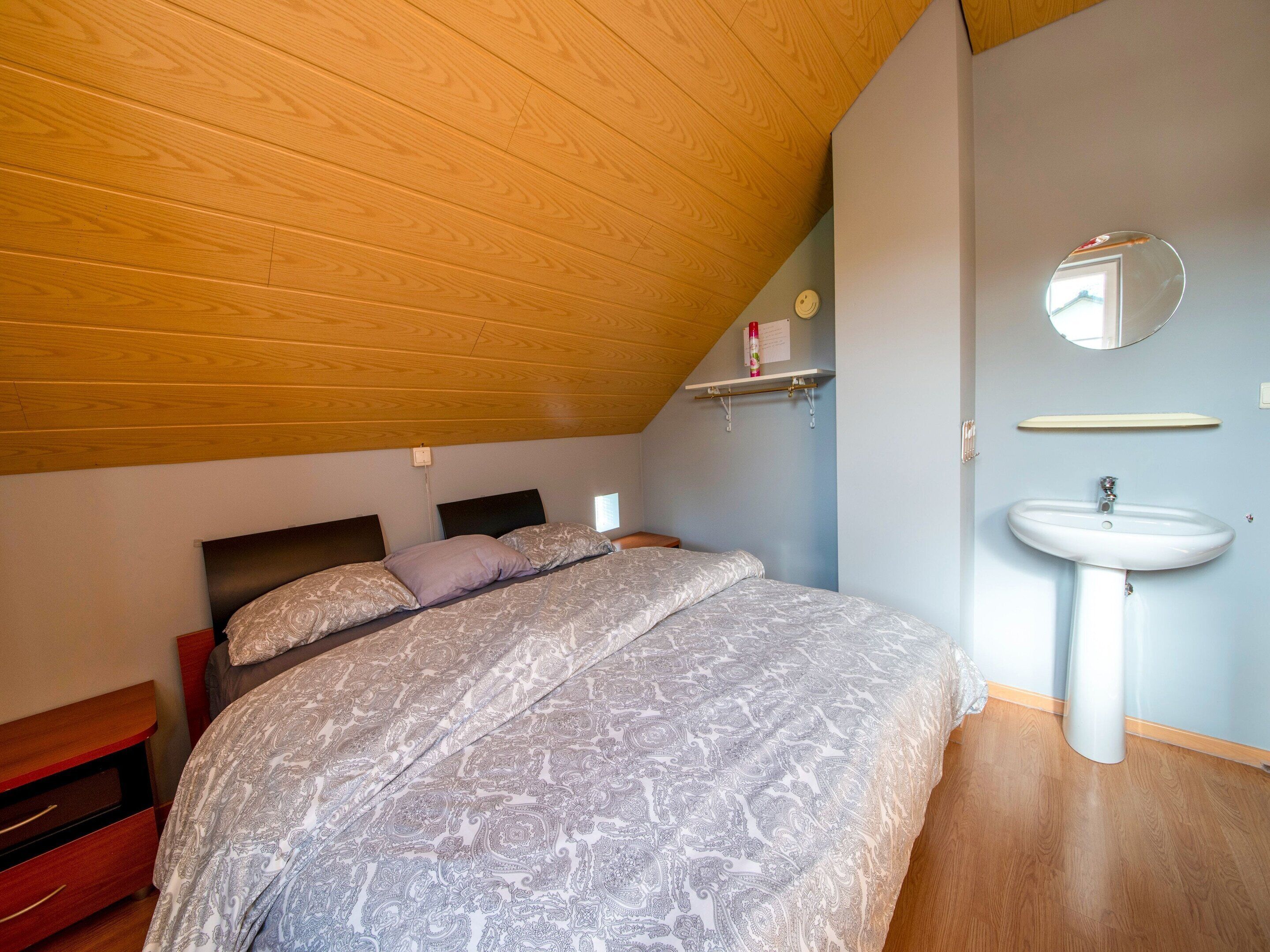 Un dormitorio con techo inclinado, una cama con ropa de cama estampada, una mesita de noche, un armario auxiliar, un estante en la pared con una botella, un espejo y un baño con lavamanos y espejo.