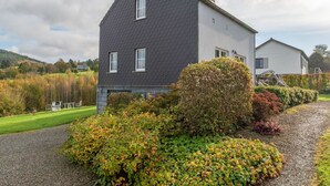 Une maison à deux étages, avec un toit sombre et des murs blancs, entourée d’une allée en gravier et d’un jardin bien entretenu, agrémenté de divers arbustes et d’une haie.