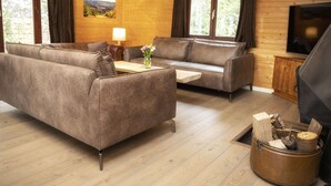 A cozy living room with a sofa, a coffee table, and a TV. There is a fireplace with logs and a wooden cabinet. - Cosy Chalet in Odeigne with Hot Tub (Manhay)