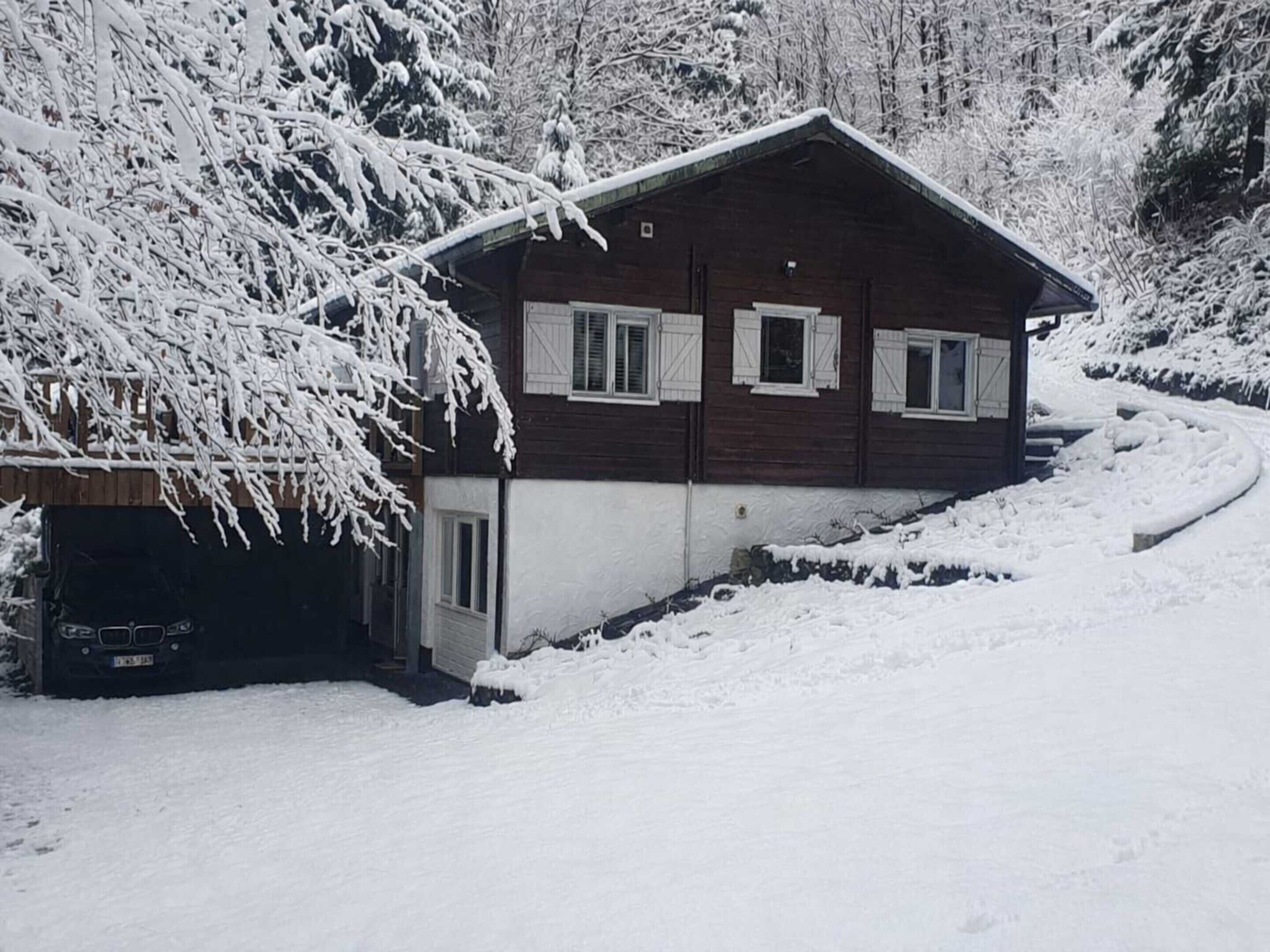 A two-story wooden cabin with white shutters, surrounded by snow-covered trees and a snow-covered driveway.