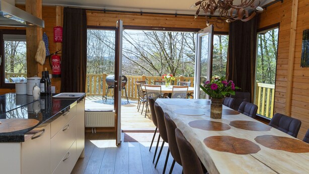 A wooden interior with a dining area, kitchen, and large windows. - Cosy Chalet in Odeigne with Hot Tub (Manhay)