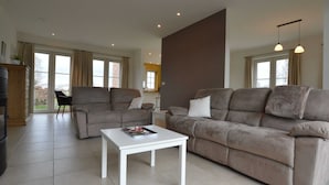 A spacious living room with a large grey sofa, a white coffee table, and a black chair. There are large windows with curtains, a wooden cabinet, and a plant on the left. - Sint-amands Country Escape (Puurs-Sint-Amands)
