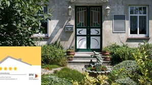 Une maison avec un toit recouvert de tuiles rouges, une porte verte et un jardin agrémenté de plantes en pot et d’une statue.