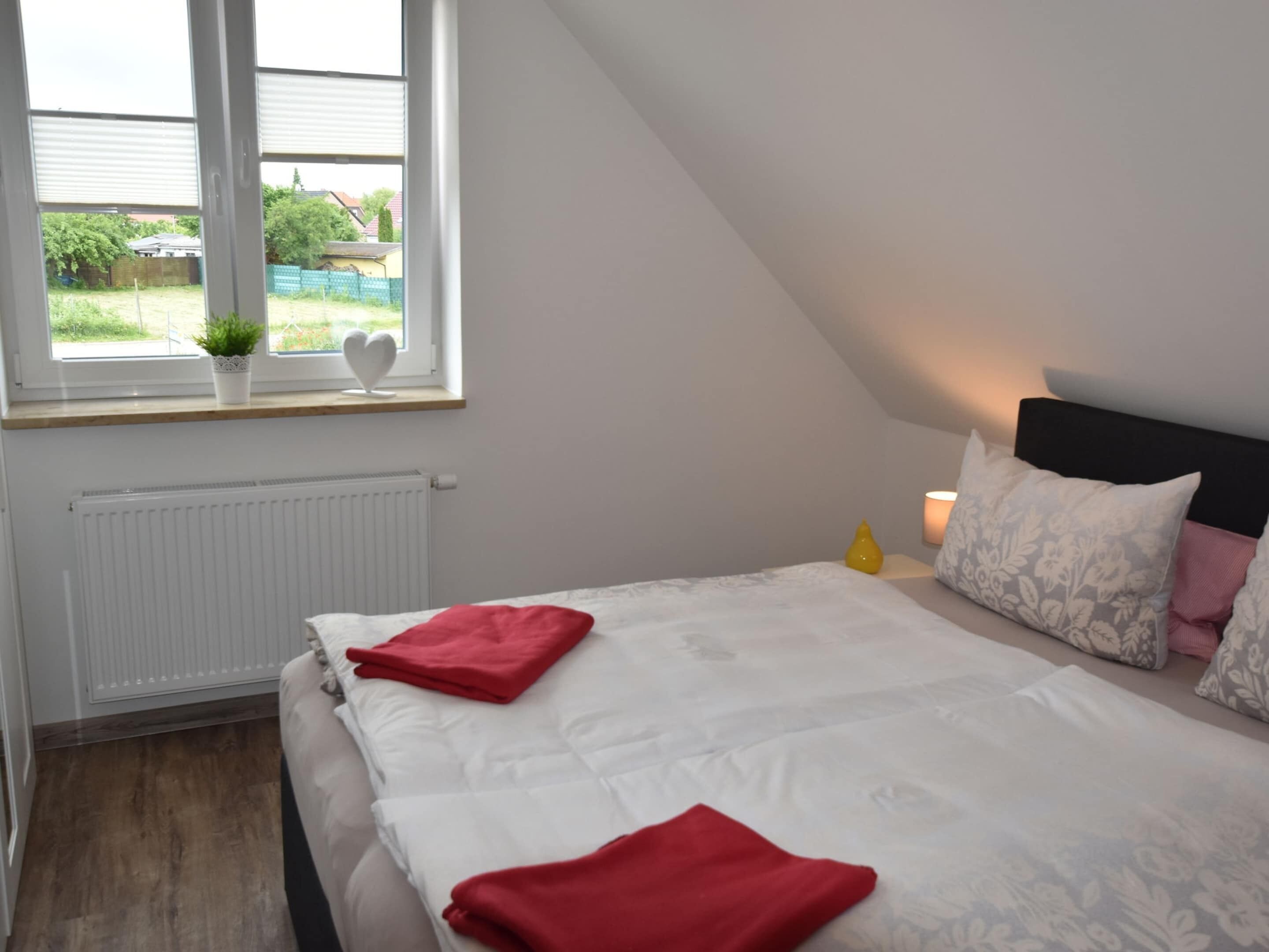 A bedroom with a bed, a window with plants, a radiator, and a bedside table with a lamp.