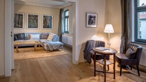 Ett mysigt vardagsrum med en soffgrupp, ett soffbord, ett sidobord med en lampa och en fåtölj. På väggen hänger inramade tavlor och det finns ett fönster med gardiner.