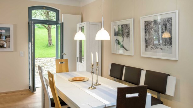 En matplats med ett vitt bord, trästolar och en bänk. Det finns två hänglampor, en dörr som leder ut och inramade tavlor på väggen.