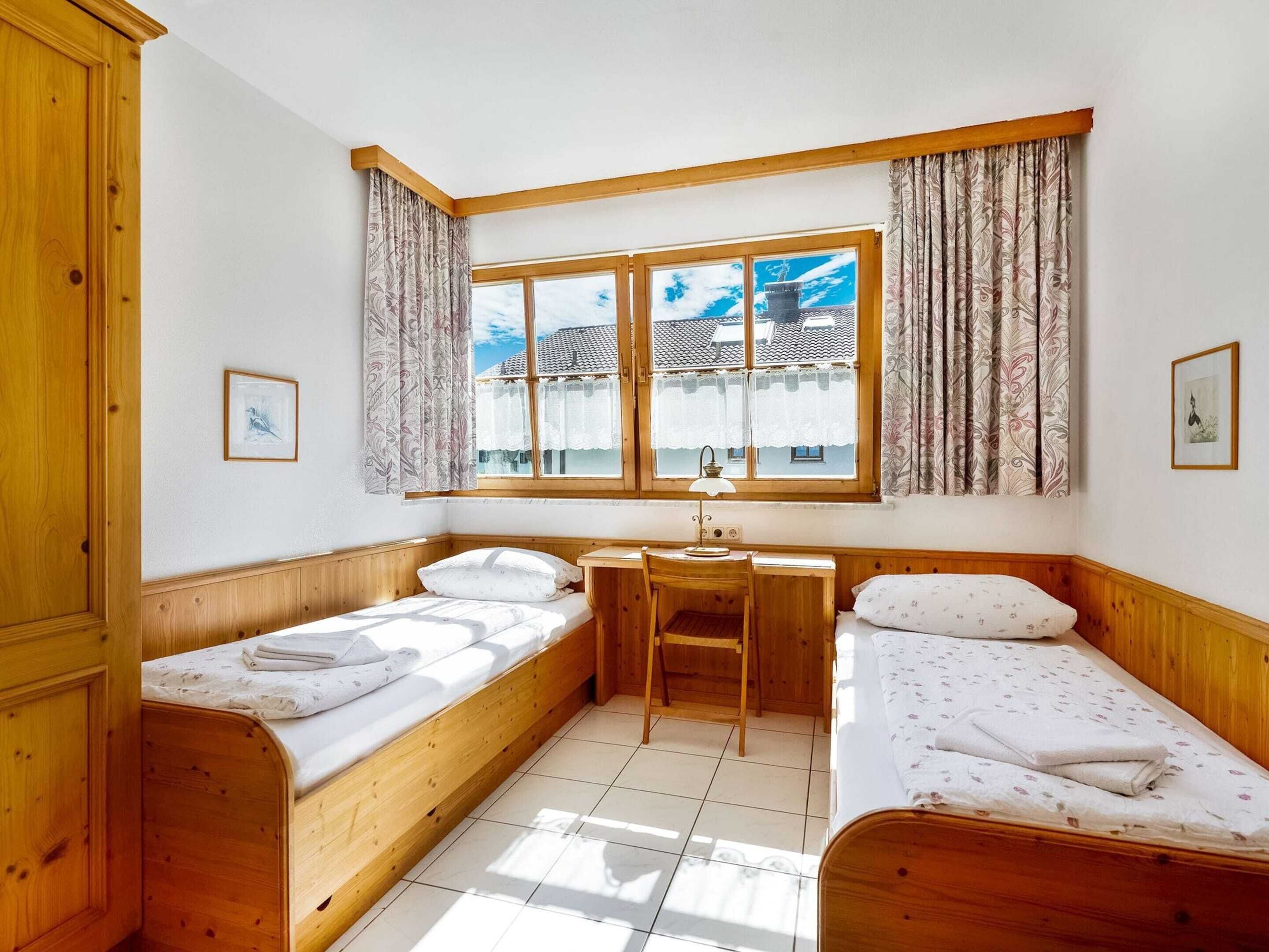 A room with two wooden beds, a desk, and a chair. There is a window with curtains, and two framed pictures on the walls.