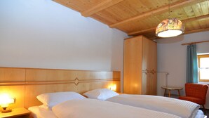 A hotel room with a wooden ceiling, two beds with white bedding, a nightstand with a lamp, a wardrobe, and a small table with a chair.