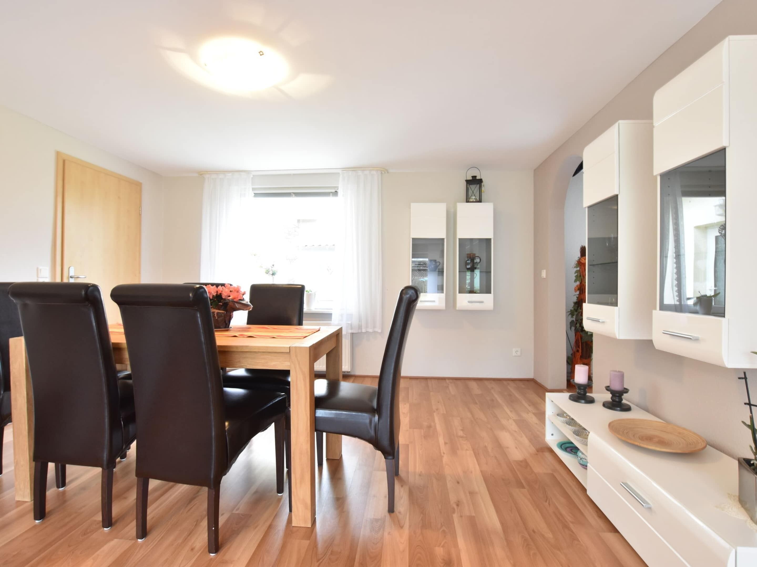 Ein Essbereich mit einem Holztisch, schwarzen Stühlen, einem weißen Schrank mit Glastüren und einer Deckenleuchte.