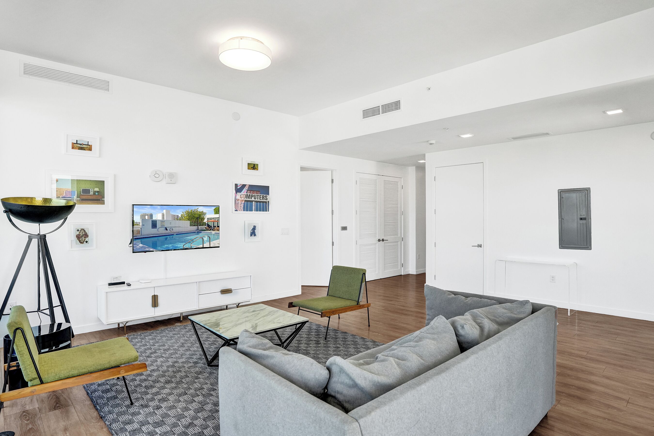 Una sala de estar moderna con un sofá gris, un sillón verde, una mesa de centro y una lámpara de pie. Hay un televisor montado en la pared y un mueble blanco debajo.