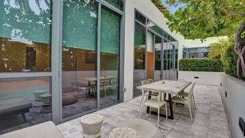 A modern outdoor patio with a dining table and chairs, a seating area with stools, and a glass door leading to an indoor space.