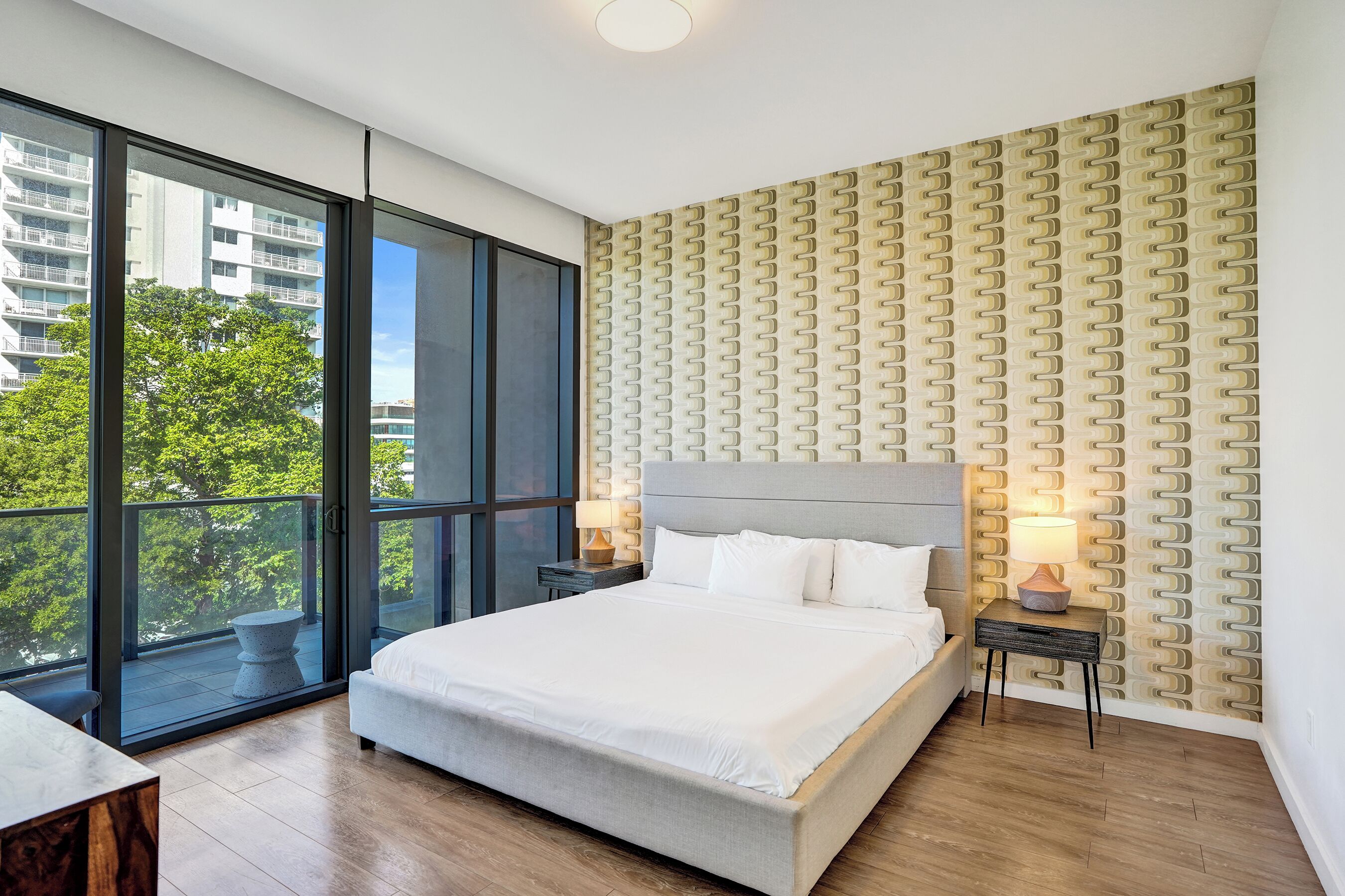 Ein modernes Hotelzimmer mit einem großen Bett, zwei Nachttischen mit Lampen, einem Balkon mit Blick auf Gebäude und Bäume sowie einer strukturierten Wand.