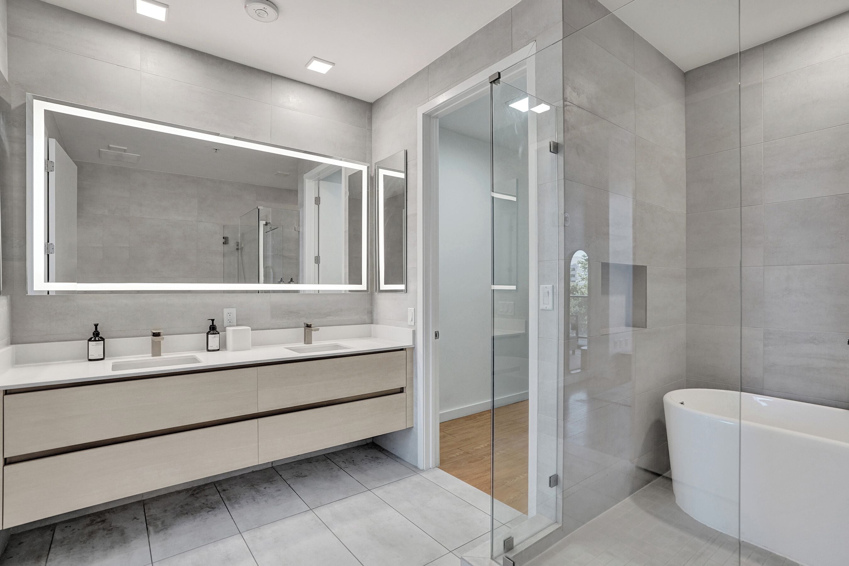 A modern bathroom with a large mirror, two sinks, a glass shower enclosure, and a bathtub.