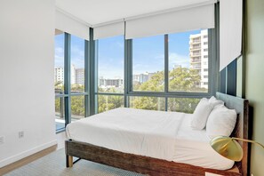 A modern bedroom with a large bed, a wooden headboard, and a view of city buildings through large windows. - 17WEST Apartments by Stay Hospitality (Miami Beach)