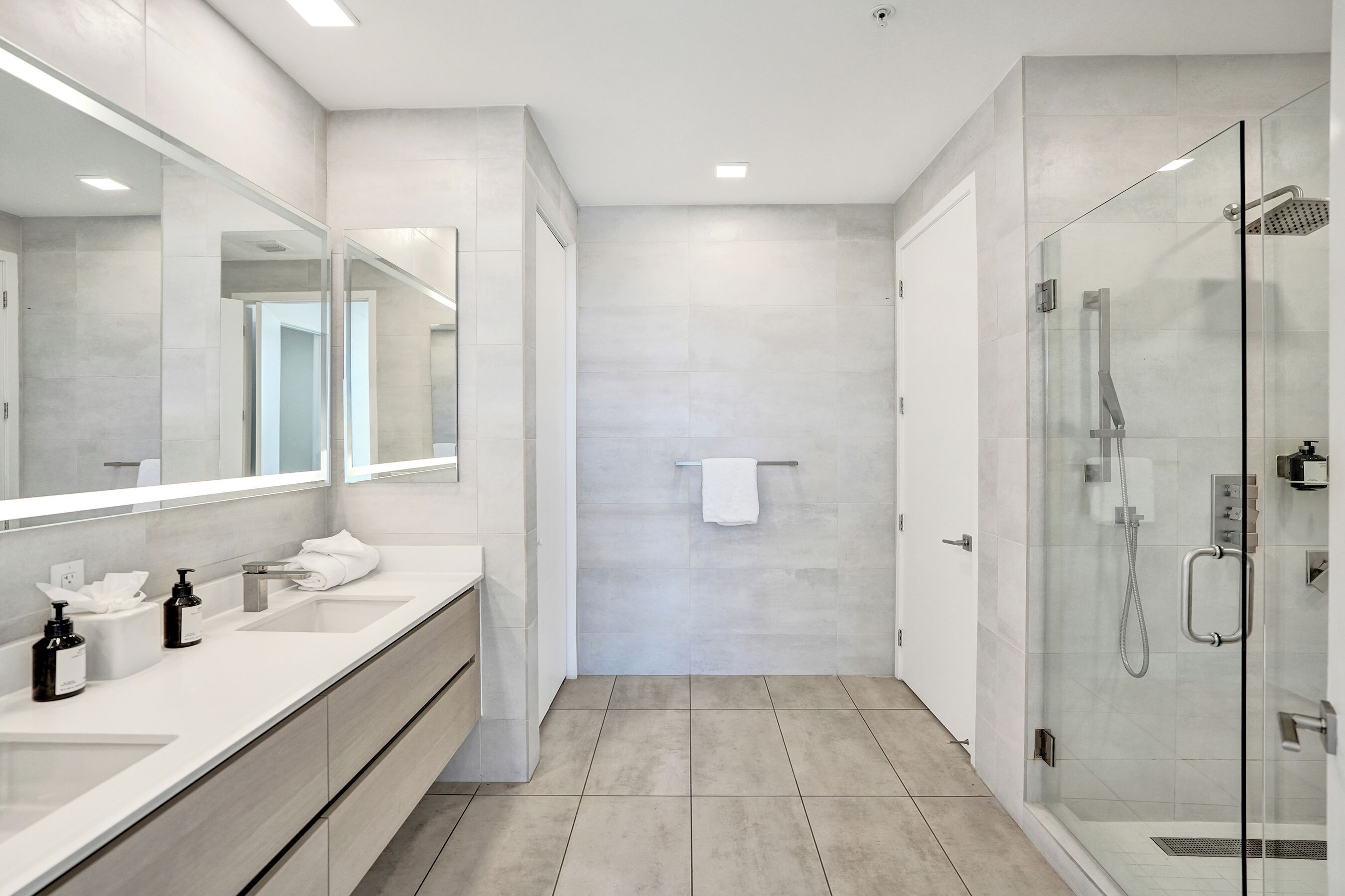 A modern bathroom with a large mirror, two sinks, a glass shower enclosure, and a towel hanging on the wall.