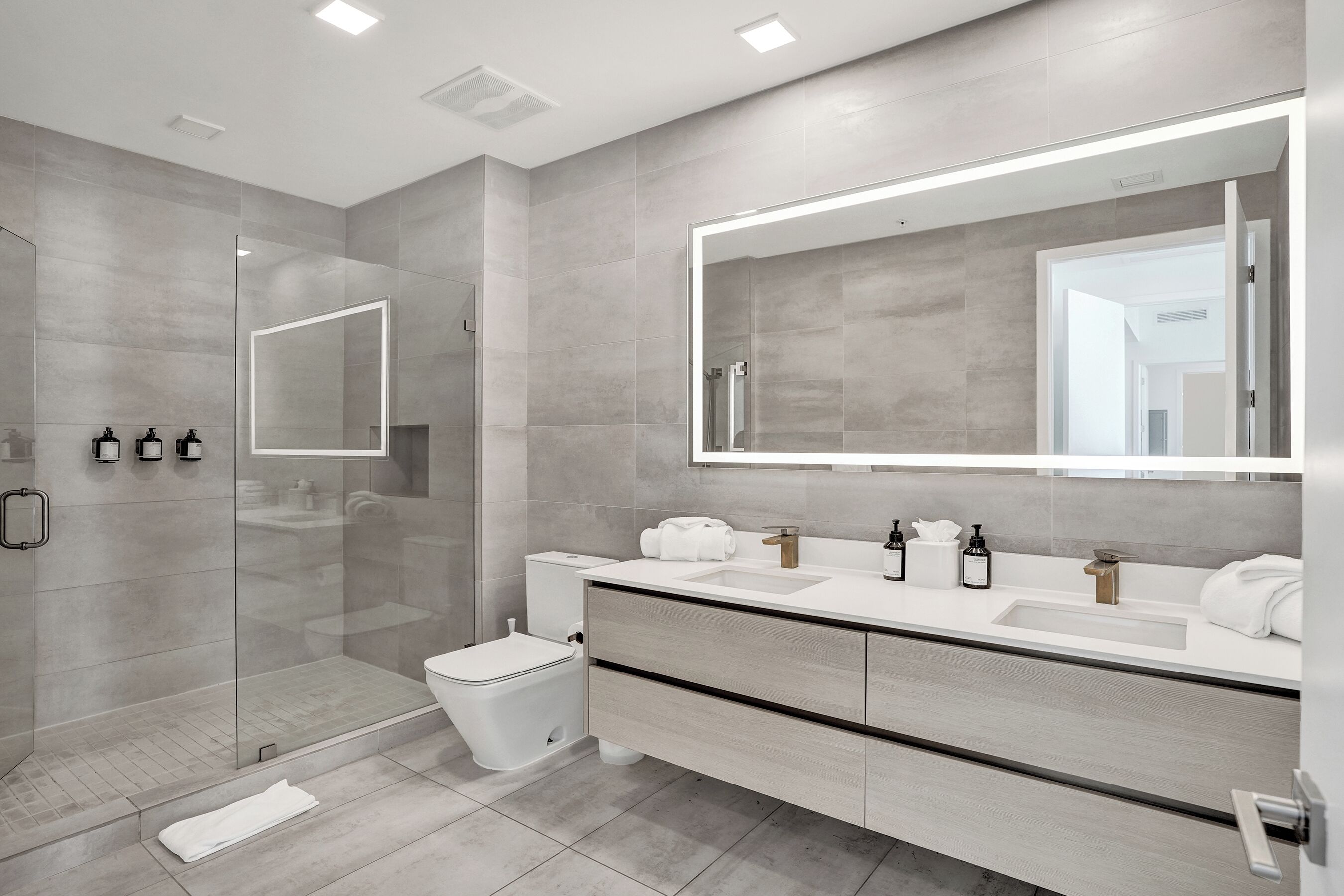 Un baño moderno con cabina de ducha de vidrio, inodoro, tocador con espejo grande y lavamanos.