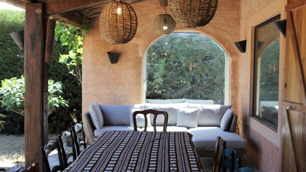 A covered outdoor seating area with a table and chairs, a wicker ceiling light, and a view through a large arched window. - Private Pool in Provence (La Motte)