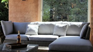 A modern outdoor seating area with a sofa and cushions, a table with a bottle, and a view of trees through a window. - Private Pool in Provence (La Motte)