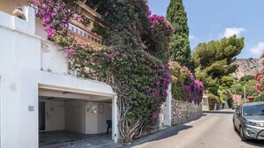 A street with a parked car, a building with a garage, and a tree with purple flowers. - Elegant Seaside Villa in Eze (Èze)