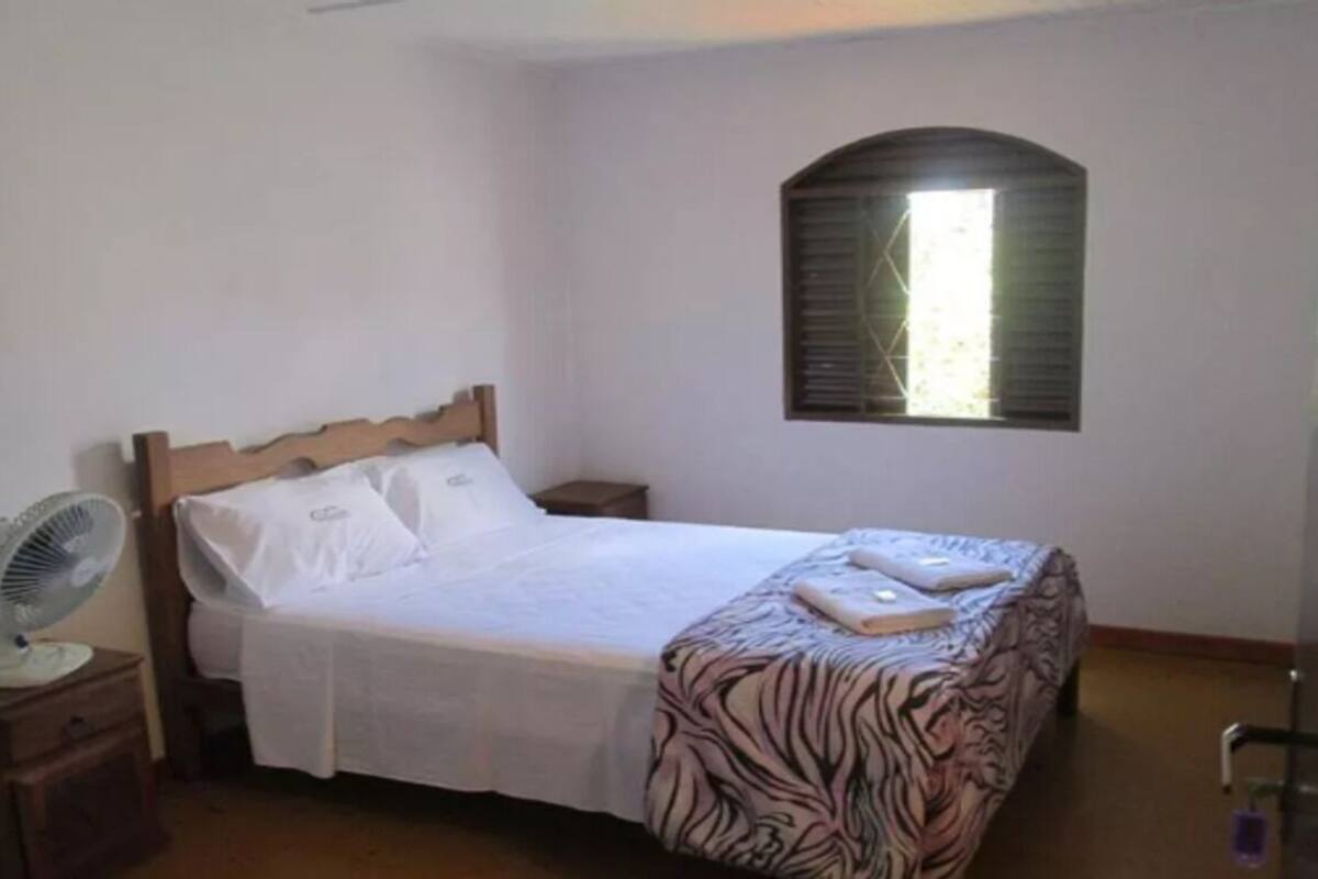 A single bed with a wooden headboard, a bedside table with a fan, and a window with shutters.