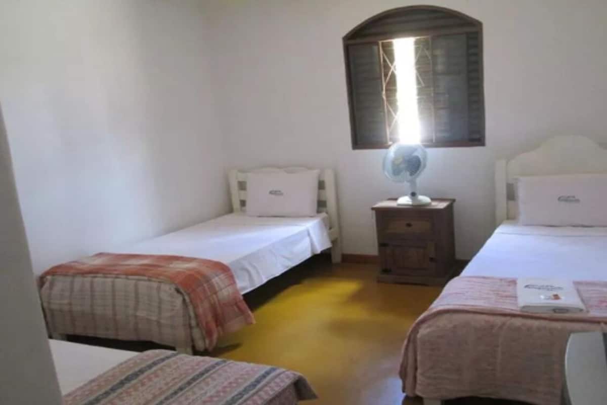 A small, simple room with two single beds, a bedside table with a fan, and a window with a wooden frame.