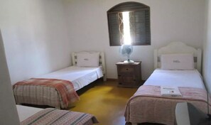 Una habitación pequeña y sencilla con dos camas individuales, una mesita de noche con un ventilador y una ventana con marco de madera.