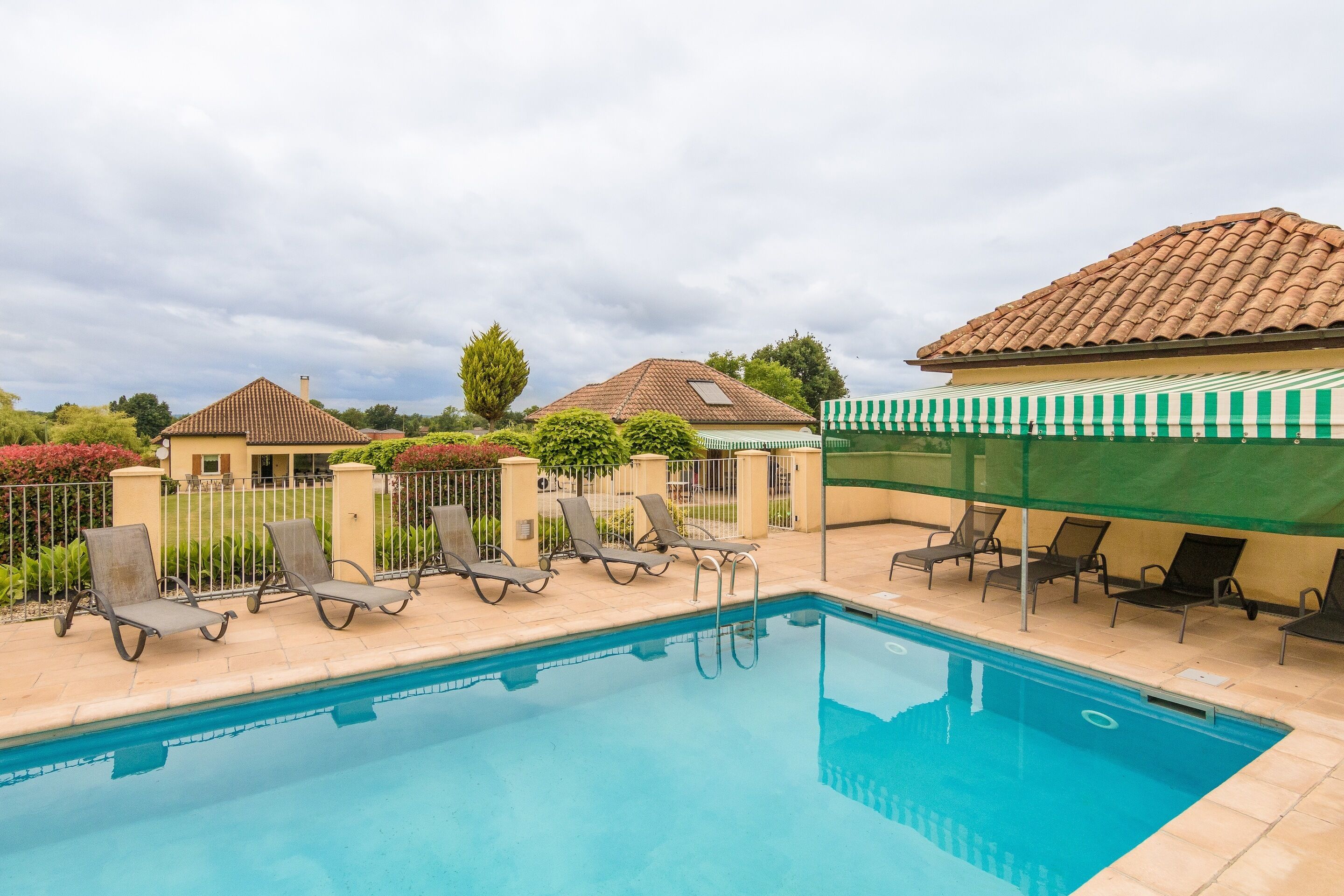 A swimming pool with lounge chairs, a covered area, and a building with a tiled roof.