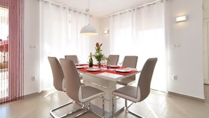 A modern dining room with a white table set for six, surrounded by six grey chairs. There is a chandelier above the table, and the room has large windows with white curtains. - Villa 1771 (Vodnjan)