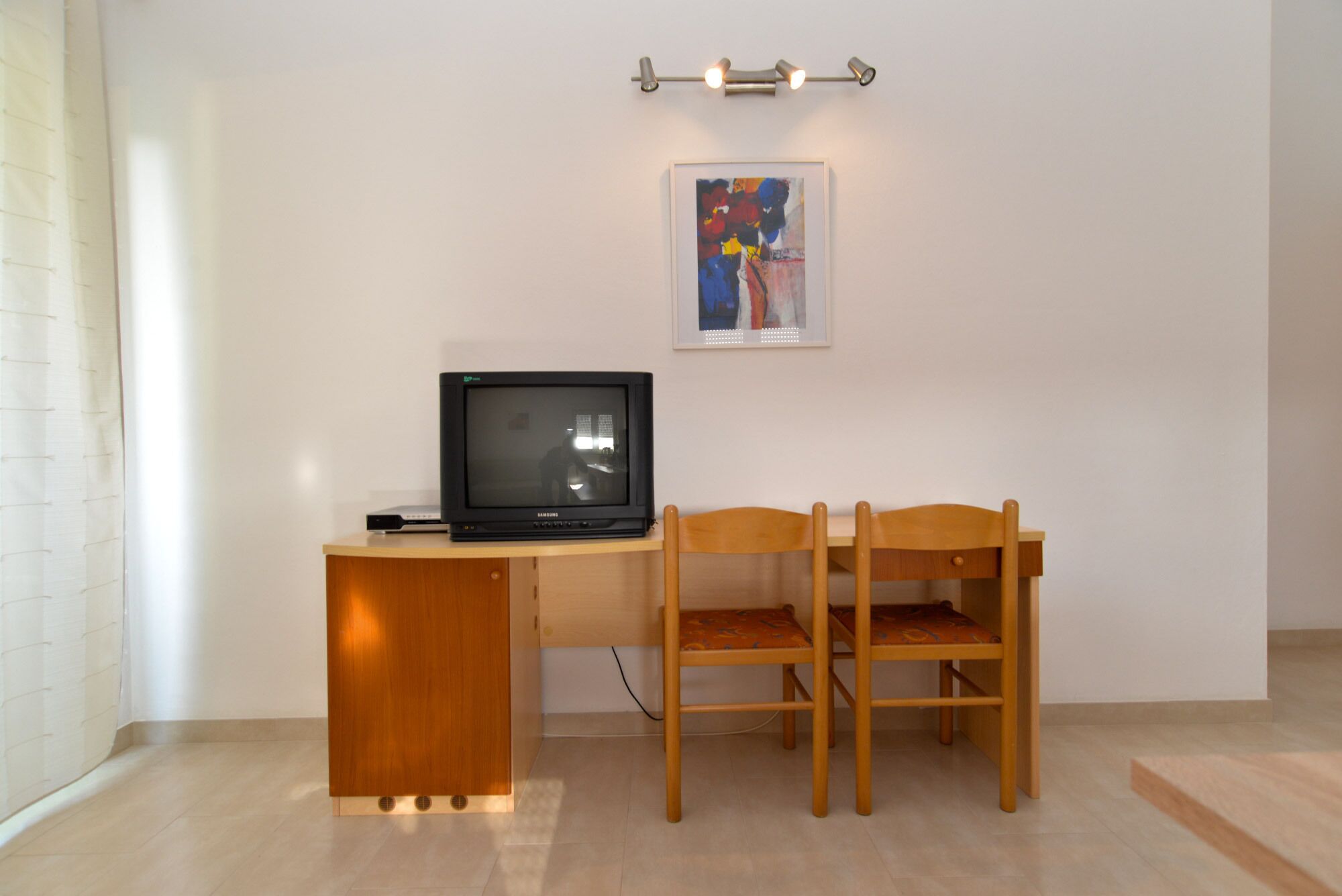 Ein kleines Zimmer mit einem Holztisch, einem Fernseher, zwei Holzstühlen und einem gerahmten Kunstwerk an der Wand.