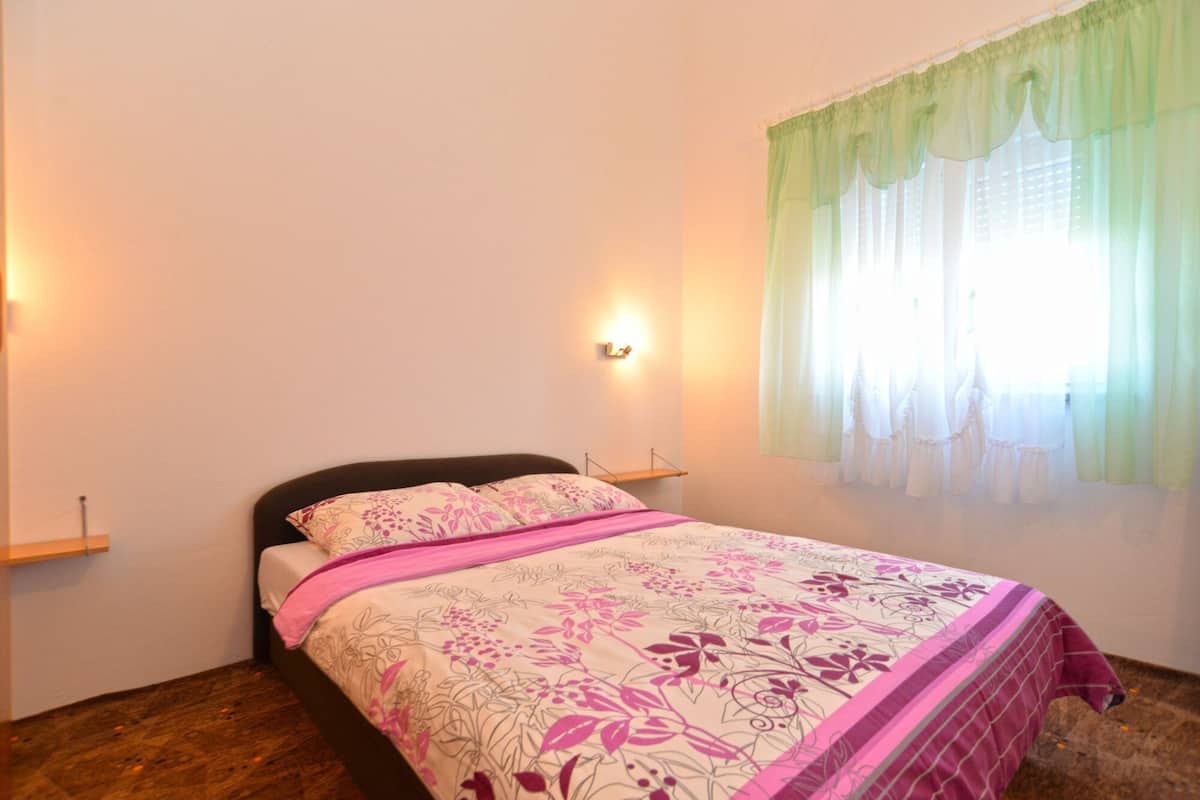 A bedroom with a bed, a nightstand, a wall-mounted light, and a window with sheer curtains.