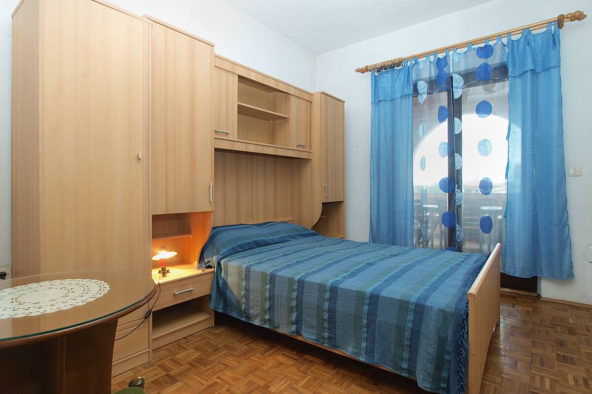Um quarto com uma cama, mesa de cabeceira, escrivaninha com abajur e guarda-roupa.