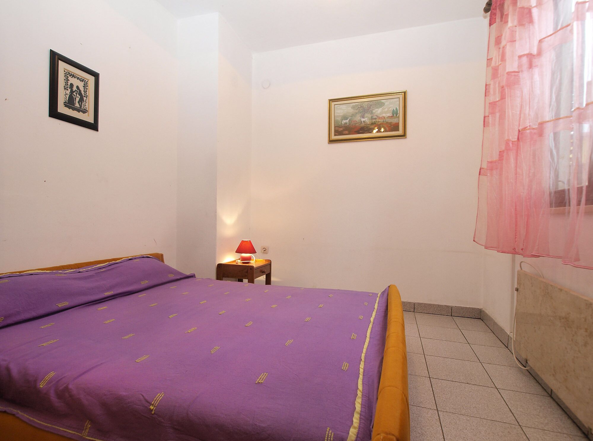Een slaapkamer met een bed, een nachtkastje, een lamp en twee foto's in lijsten aan de muur.