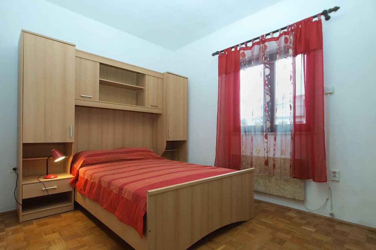 Quarto com cama de madeira, escrivaninha com abajur e janela com cortinas vermelhas.
